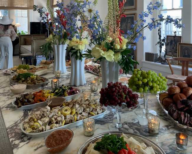 A large table is adorned with an extensive spread of food, including various fruits, vegetables, and dishes, surrounded by vases of fresh flowers and softly glowing candles.