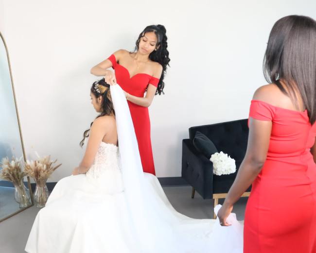 A woman in a white wedding dress sits in front of a mirror while another woman adjusts her veil and a third woman stands nearby, both wearing red dresses.