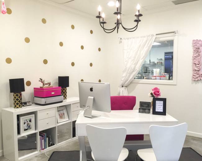 The image shows a modern office space with a white desk, a stylish computer, a pink chair, decorative light fixtures, and gold polka dots on the wall.