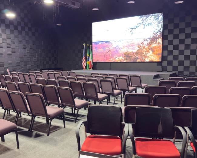 The image shows a conference room with rows of chairs facing a large screen displaying a landscape, flanked by two flags.