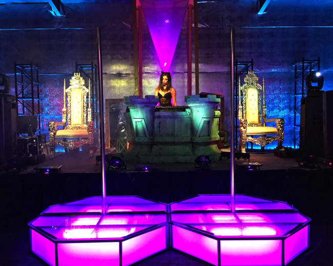 A DJ performs on stage in front of ornate thrones, surrounded by colorful lighting and large speakers.