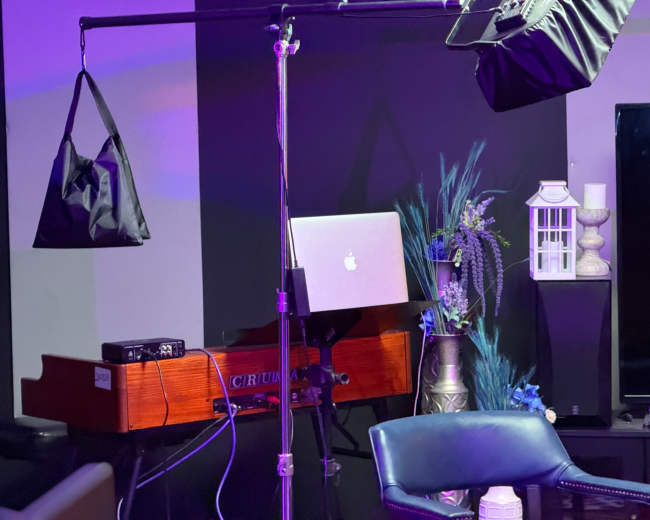 The image shows a music setup featuring a keyboard, a laptop on a stand, a light fixture, and decorative plants on display in a studio-like environment.