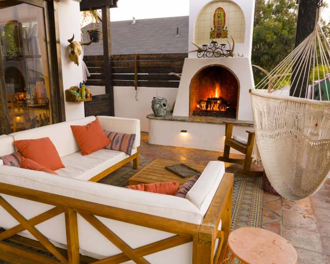 The image shows a cozy outdoor seating area featuring a white sectional couch with orange cushions, a wooden coffee table, and a hammock next to a built-in fireplace.