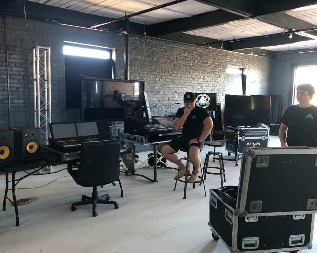 A music production studio with multiple computers, audio equipment, and two individuals, one seated and the other standing.