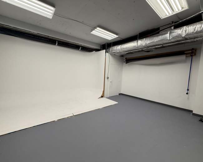 The image shows a bare photography studio with a white backdrop, grey floor, and exposed ventilation ducts.
