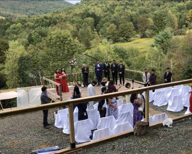 A wedding ceremony takes place outdoors on a deck overlooking a green landscape, with guests seated in rows and a floral arch at the front.