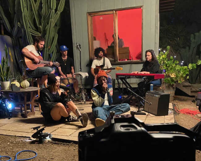 A group of musicians performs together in a backyard setting, with instruments and equipment visible, while some sit on the ground in front of a lit window.