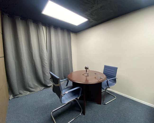 The image shows a small, sparsely furnished room with a round table and two chairs, surrounded by grey curtains and a ceiling light.