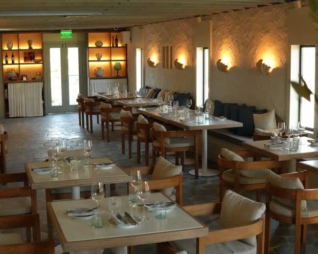 The image shows a modern restaurant interior with wooden tables, chairs, and soft lighting, featuring neatly set dining arrangements.
