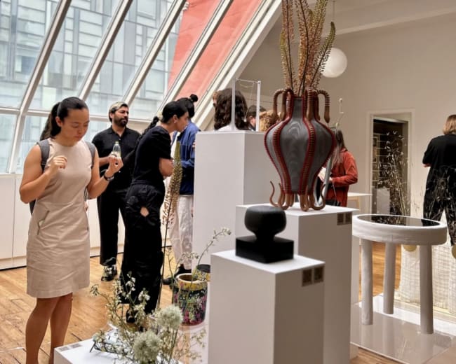 The image shows a group of people interacting with various art pieces displayed on white pedestals in a bright, modern gallery with large windows.
