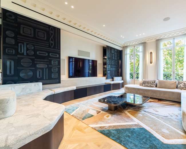 The image shows a modern living room featuring a curved sofa, a large television mounted on a dark wall, and ample natural light from large windows.