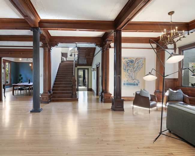 The image shows a spacious interior with wooden pillars, a staircase in the background, and a combination of modern and traditional furniture.