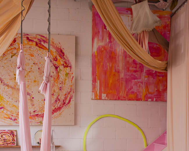 The image shows a cozy, brightly colored living space with pink fabric draped from the ceiling, a circular art piece on the wall, and a seating area featuring a light brown sofa and a small table.