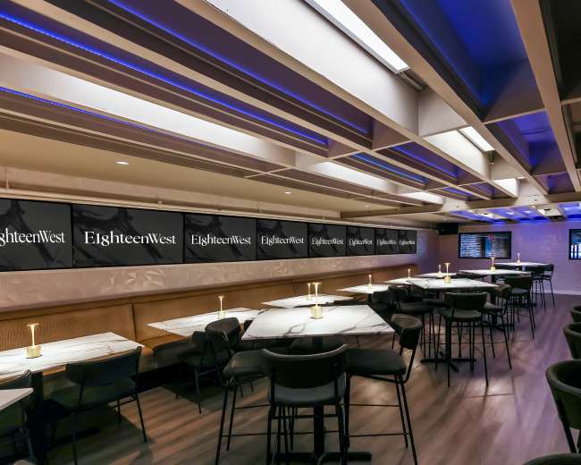 The image shows a modern dining area with marble-topped tables and black chairs, featuring ambient lighting and a large wall display with the name "EighteenWest."