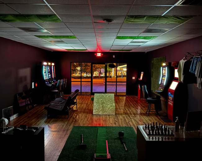 The image depicts a dimly lit entertainment space with several arcade machines, a golf putting area, and a large window showing an outdoor view at night.