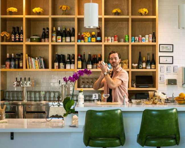 A bartender shakes a cocktail behind a well-stocked bar featuring various bottles and decorative yellow flowers.