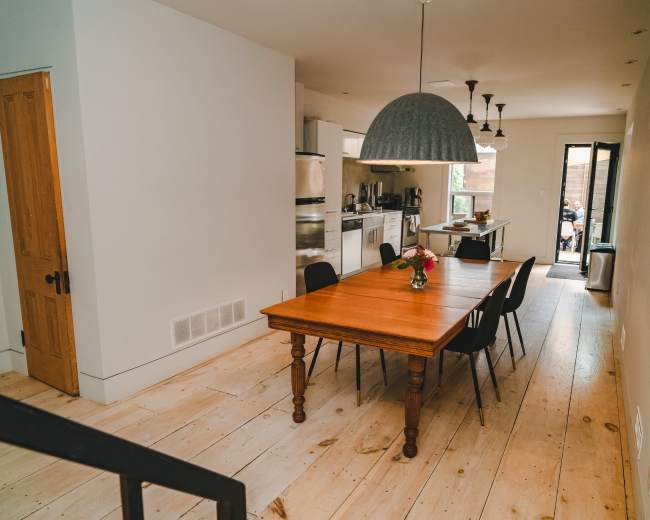 A modern kitchen and dining area features a wooden dining table surrounded by black chairs, with a large pendant light overhead and an open view of the kitchen space.
