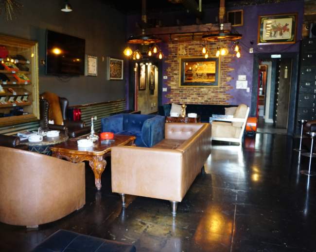 The image shows a stylish lounge area featuring brown leather sofas, a blue velvet couch, wooden tables, and decorative lighting against a dark-themed interior.