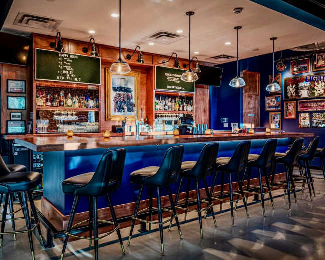 A modern bar features a long wooden counter with high stools, illuminated by pendant lights, surrounded by shelves filled with various bottles and framed artworks on the walls.