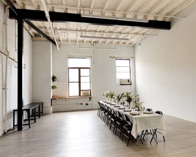 A long dining table is set up in a spacious, minimalist room with large windows allowing natural light to fill the space.
