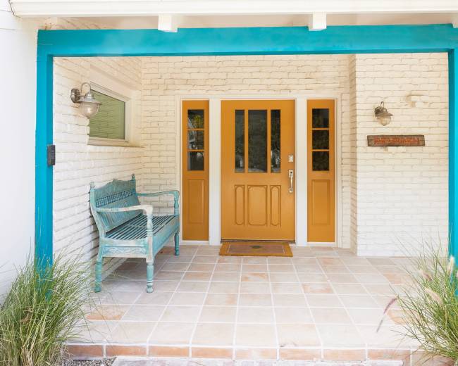 The image shows a front porch with a turquoise trim, a yellow double door, a decorative bench, and potted plants.