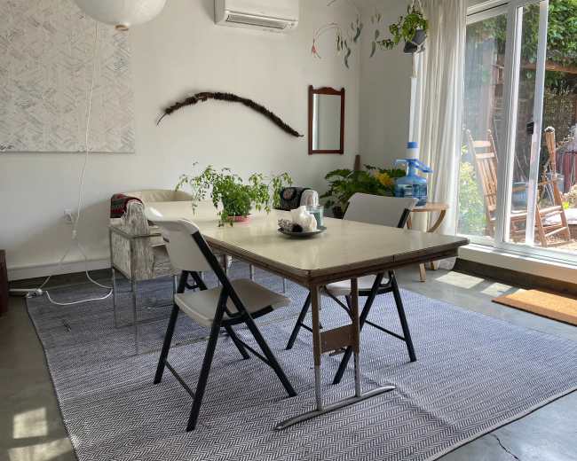 A dining area features a table surrounded by chairs, with plants, a decorative wall hanging, and sliding glass doors leading to an outdoor space.