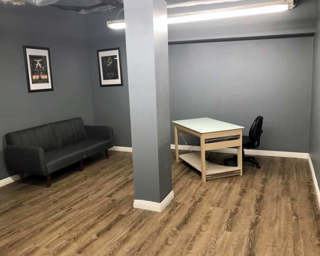 The image shows an empty room with gray walls, a wooden floor, a gray couch against one wall, a desk with a chair in the center, and two framed posters on the walls.