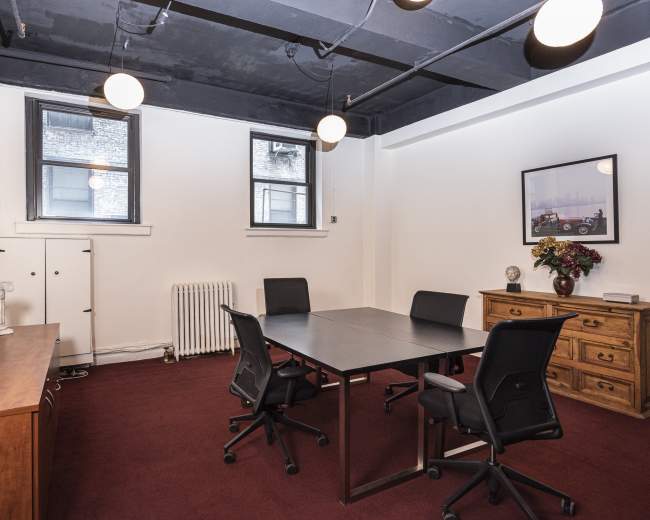 A conference room features a rectangular table surrounded by four chairs, with windows, a cabinet, and a decorative photo on the wall.