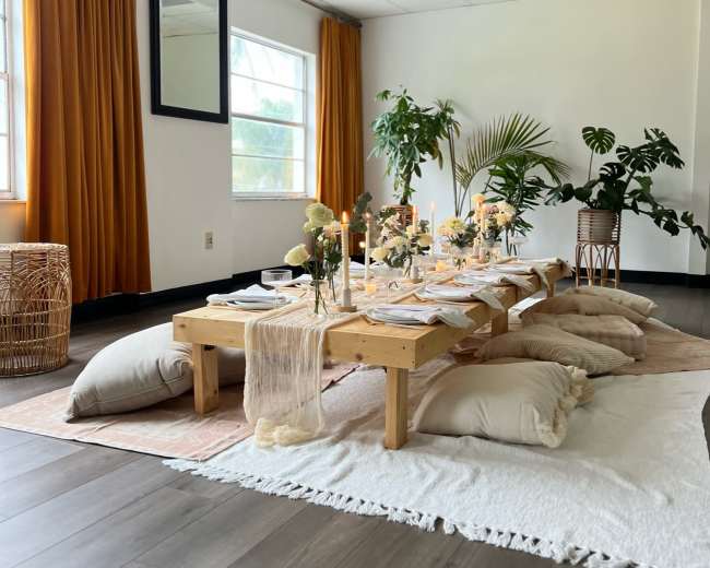 A dining area features a low wooden table set with dishes and floral centerpieces, surrounded by cushions on a decorative rug.