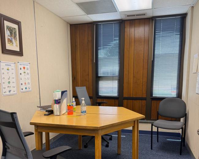 The image shows a small office with a wooden hexagonal table, two chairs, a water bottle, and a small assortment of office supplies.