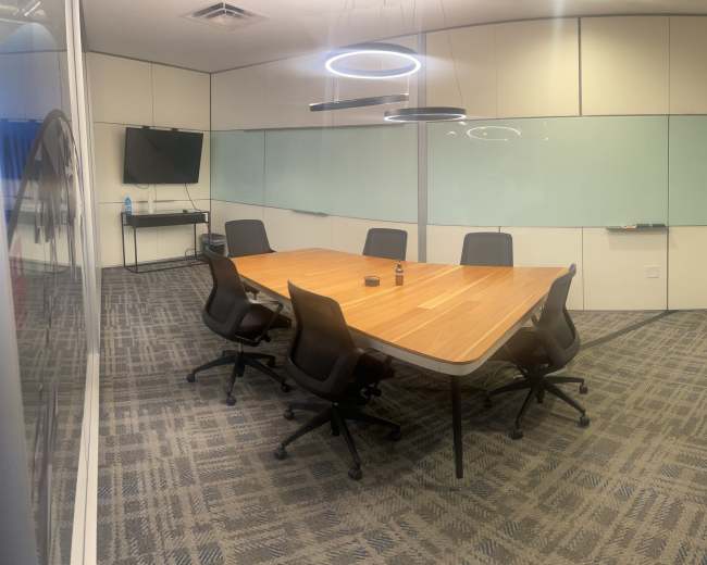 A modern conference room features a large wooden table surrounded by six ergonomic chairs, with a television and glass walls.