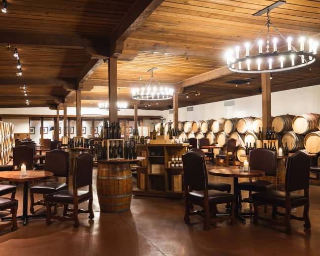 The interior of a wine tasting room features wooden barrels lining the walls, circular chandeliers overhead, and tables with chairs arranged throughout the space.