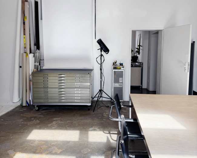 The image shows a bright, minimalistic meeting room with a large table, black chairs, a projector screen, and storage cabinets.