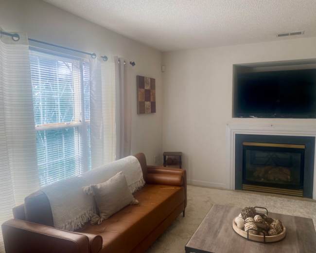A living room features a brown leather couch, a wooden coffee table with decorative items, and a wall-mounted television above a fireplace.