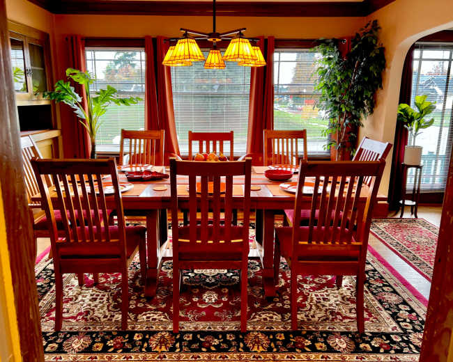 A dining room features a wooden table set for eight, surrounded by matching chairs, with decorative plants and large windows allowing natural light.