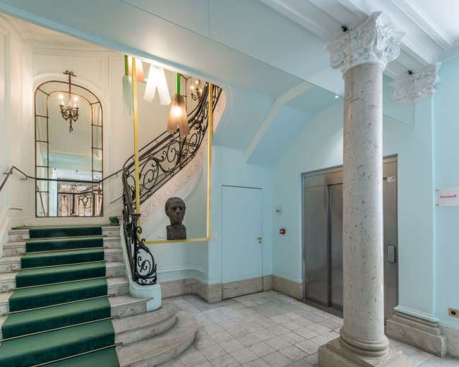 The image shows a well-lit interior with a staircase featuring a wrought-iron railing, a marble column, and a bust on a pedestal.