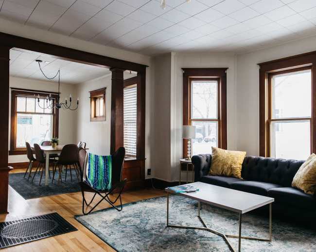 A living room with a dark sofa, a coffee table, and a chair, adjacent to a dining area with a table and chairs, illuminated by natural light through large windows.