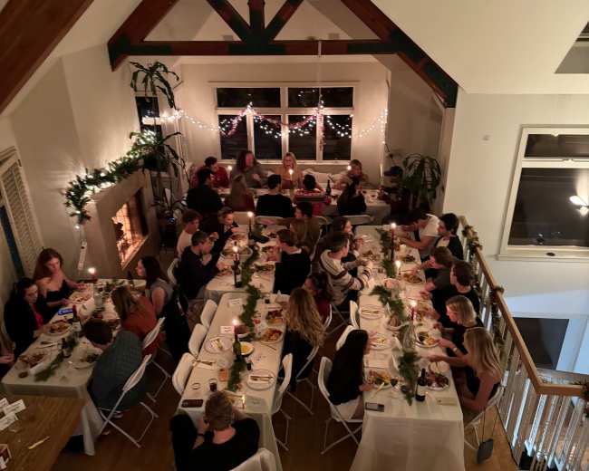 A large group of people is gathered around several long tables set for a dinner in a warmly lit room with wooden beams and decorative greenery.