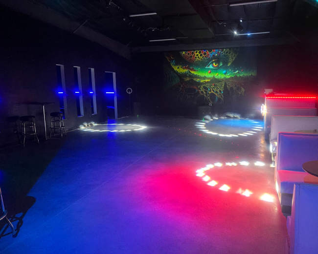 The image shows a dark, spacious venue with a colorful mural on the wall and various circular light patterns projected on the floor.