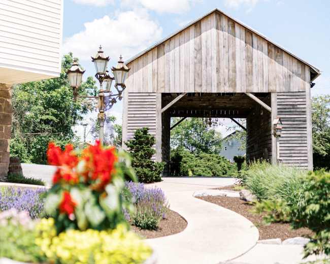 A stone pathway leads through landscaped gardens to a wooden structure under a blue sky with clouds.