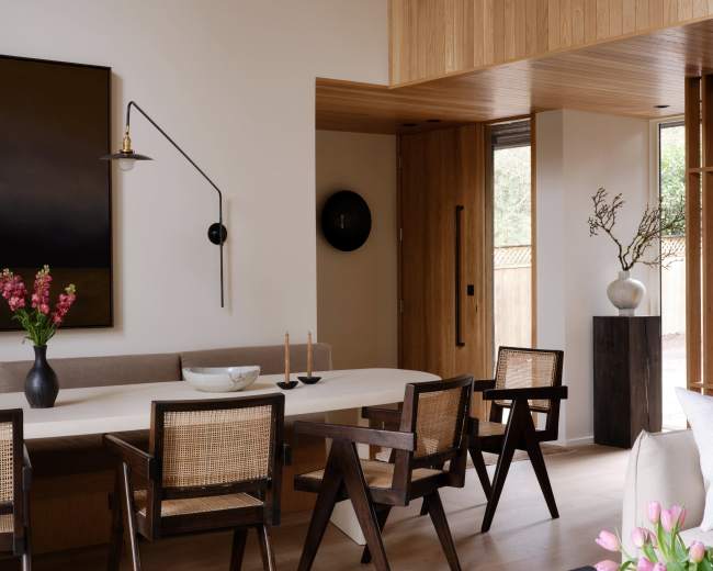 A modern dining area features a long, light-colored table surrounded by wooden chairs, with a large painting on the wall and decorative elements on display.