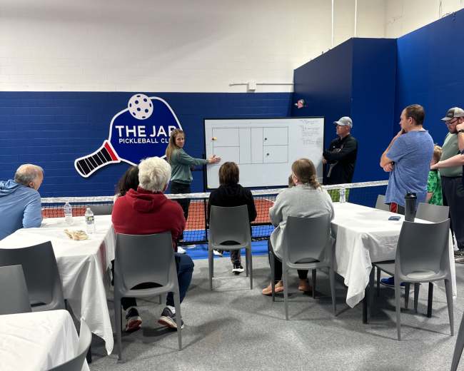 A group of people is gathered in a indoor pickleball facility, listening to a presentation near a pickleball court.