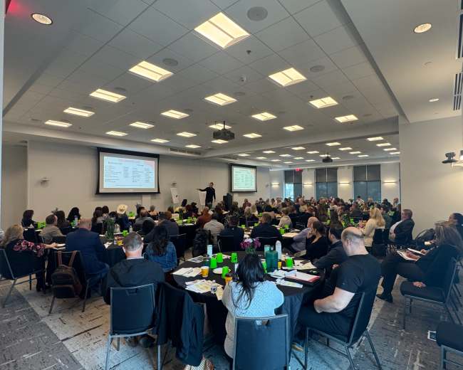 A large group of people sits at tables in a conference room, attentively watching a presenter speaking at the front of the room.
