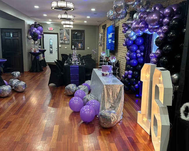 A decorated event space featuring a sparkling silver table, black and purple balloons, and a large illuminated "16" sign.