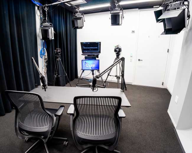 The image shows a video production studio equipped with two chairs, a large table, and multiple mounted lights and cameras.