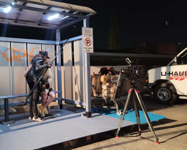 A film crew sets up equipment at a bus stop shelter under artificial lights in an urban area at night.