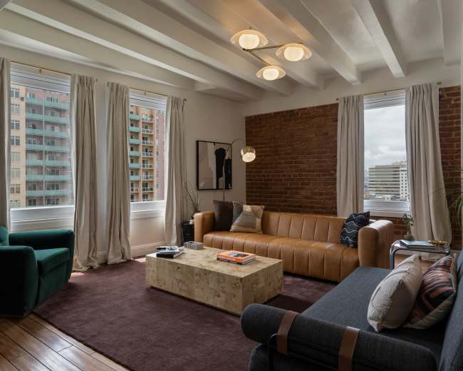 The living room features a leather sofa, a coffee table made of light-colored stone, a gray sofa, and large windows with sheer curtains overlooking a cityscape.