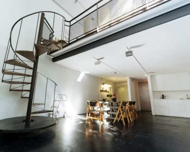 The image shows a modern, open-concept interior featuring a spiral staircase, wooden dining chairs, and a kitchen area in the background.