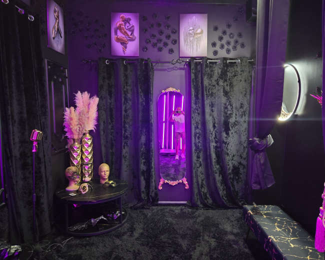The image shows a dark, elegantly decorated room with black curtains, a large mirror framed in gold, and purple lighting creating a moody atmosphere.