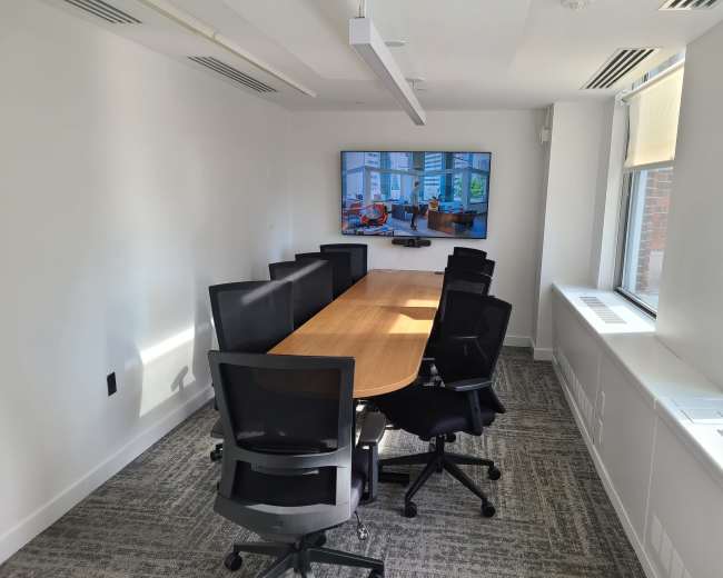 A modern conference room features a long wooden table surrounded by black ergonomic chairs, with a large screen mounted on the wall displaying a bright, airy interior.